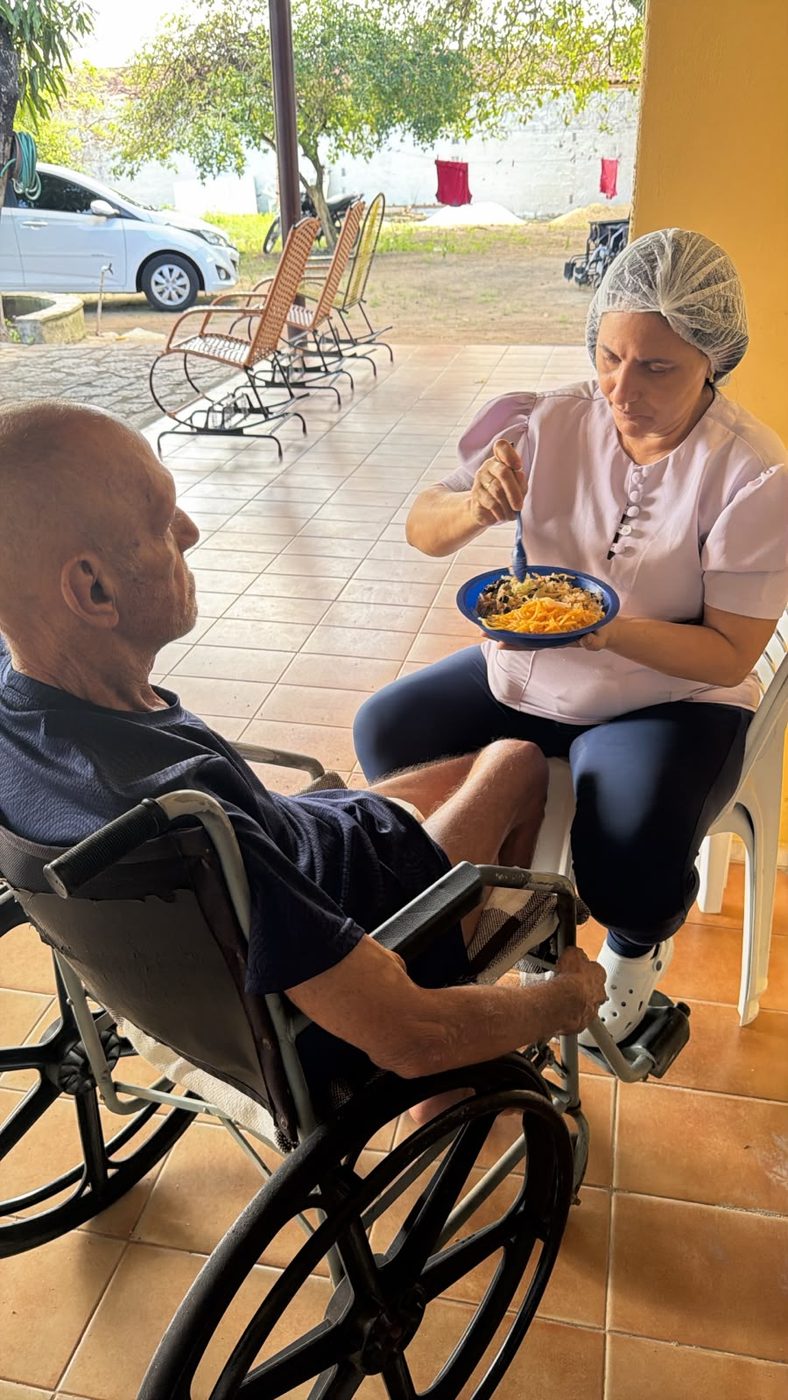 Cuidadora de touca e uniforme claro serve uma refeição em um prato azul a um morador em cadeira de rodas, na varanda ensolarada do lar