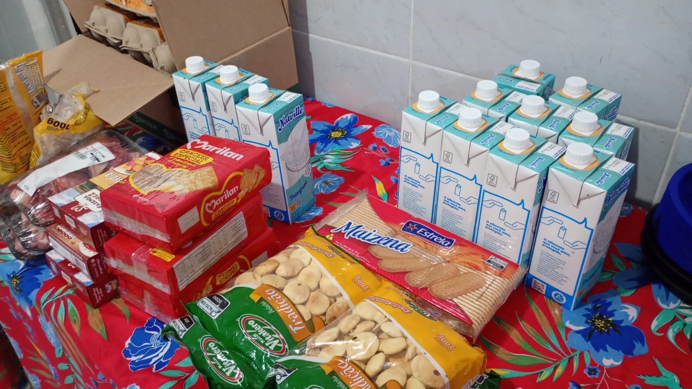 Mesa com toalha florida vermelha cheia de caixas de leite, pacotes de biscoito e outros alimentos doados ao lar