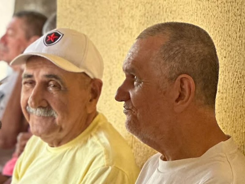 Dois moradores sentados lado a lado em uma varanda de parede amarela; o de boné branco e bigode sorri para a câmera