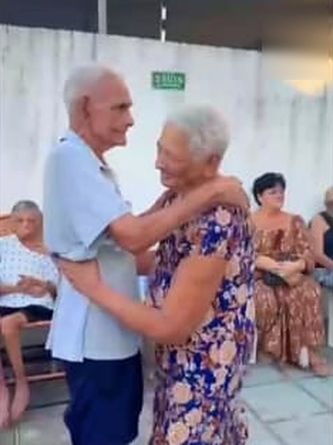 Um idoso de camisa azul-clara e uma idosa de vestido florido se abraçam sorrindo em um pátio, com outros moradores sentados ao fundo