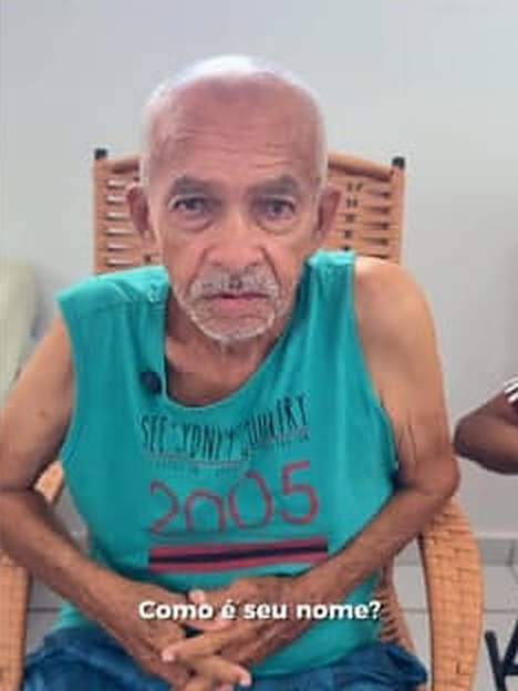 Morador de camiseta verde-água, sentado em uma cadeira de vime, olha para a câmera; a legenda do vídeo pergunta: Como é seu nome?