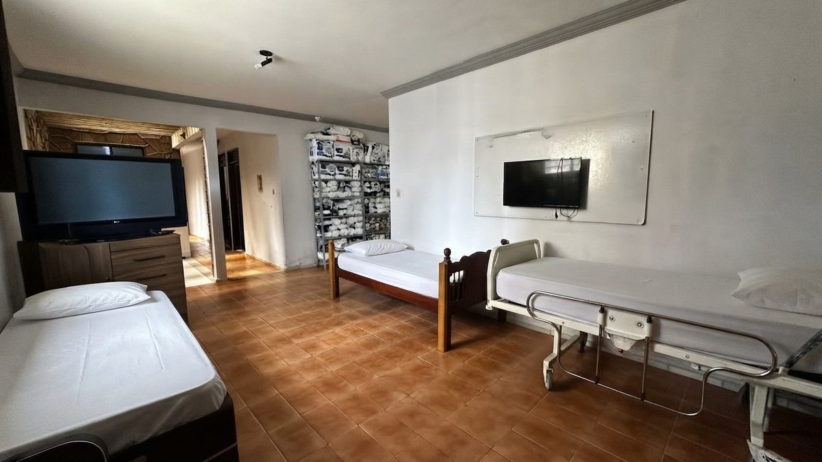 Ambiente com camas de solteiro e uma cama hospitalar, televisão na parede e prateleira com suprimentos