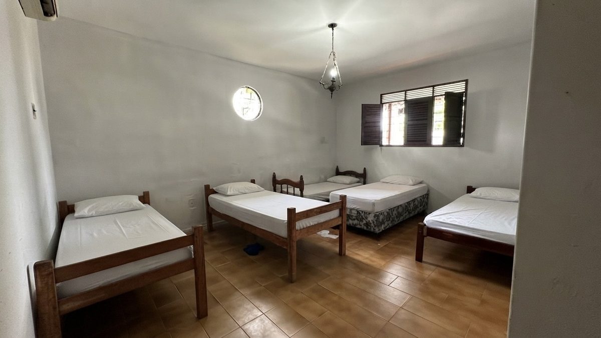 Dormitório amplo com várias camas de solteiro arrumadas, janela redonda e lustre no teto