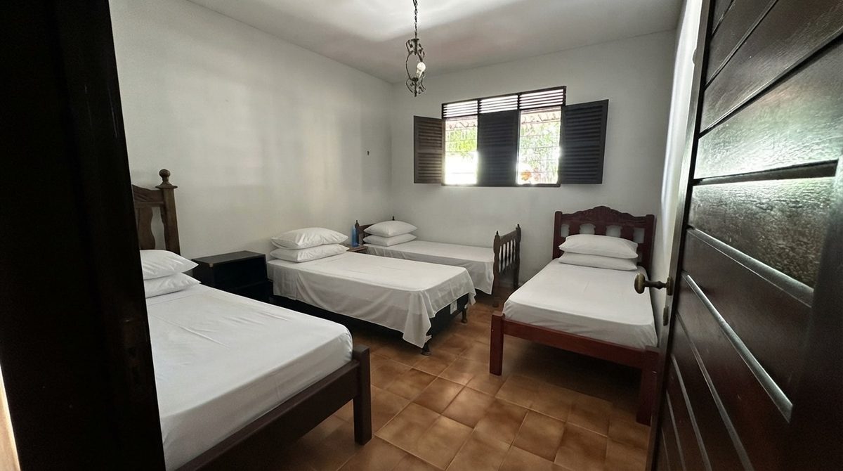 Dormitório com três camas de cabeceira de madeira escura, travesseiros brancos e mesa de cabeceira