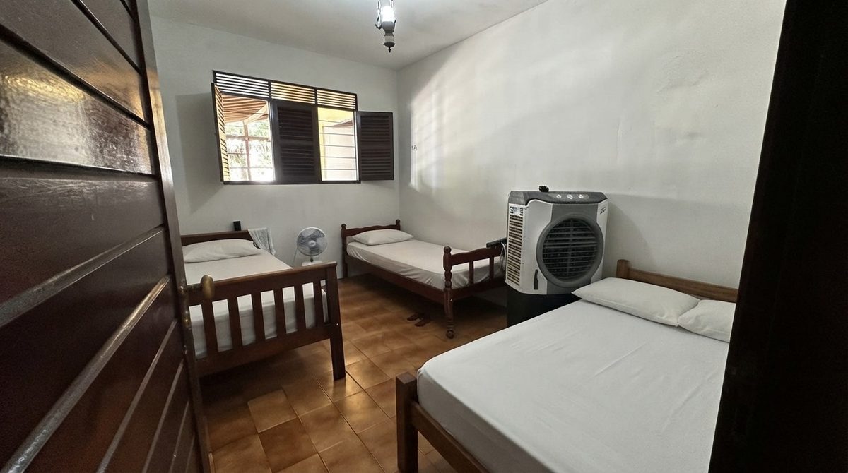 Dormitório com camas arrumadas, ventilador e climatizador de ar ao fundo e janela com venezianas