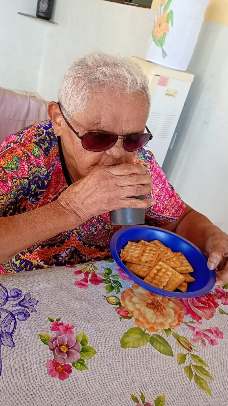 Moradora de cabelos brancos, óculos escuros e blusa colorida bebe de um copo à mesa, com um prato de biscoitos ao lado