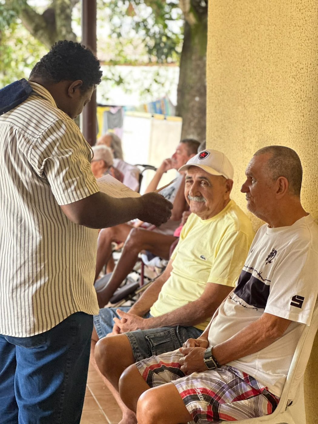 Morador de boné branco e camiseta amarela sorri para a câmera ao lado de outro morador, em uma varanda de parede amarela