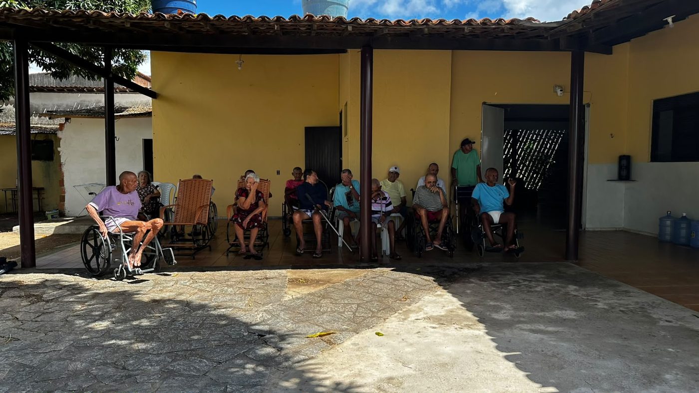 Pátio coberto do lar, com paredes amarelas e moradores sentados em cadeiras de rodas e de balanço, em um dia de sol